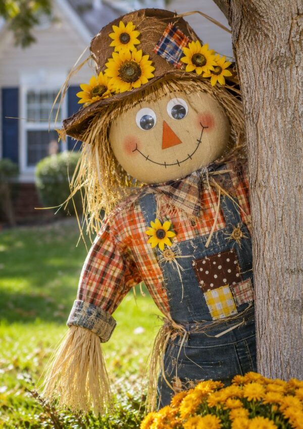 A cheerful scarecrow with a plaid shirt, overalls, and a sunflower hat leans against a tree in a sunny yard, smiling with painted features. Yellow flowers bloom at its base.