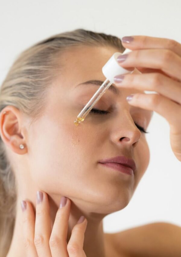 A woman with closed eyes applies facial serum to her cheek using a dropper, gently touching her neck with her other hand.