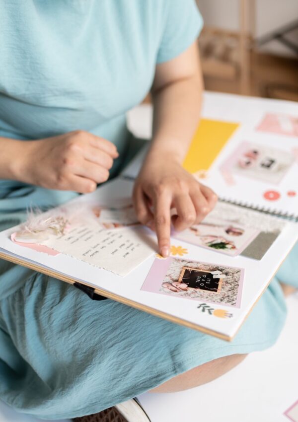 A person in a light blue dress sits cross-legged, pointing at photos and decorative elements in an open scrapbook. The book contains pictures, handwritten notes, and colorful accents.