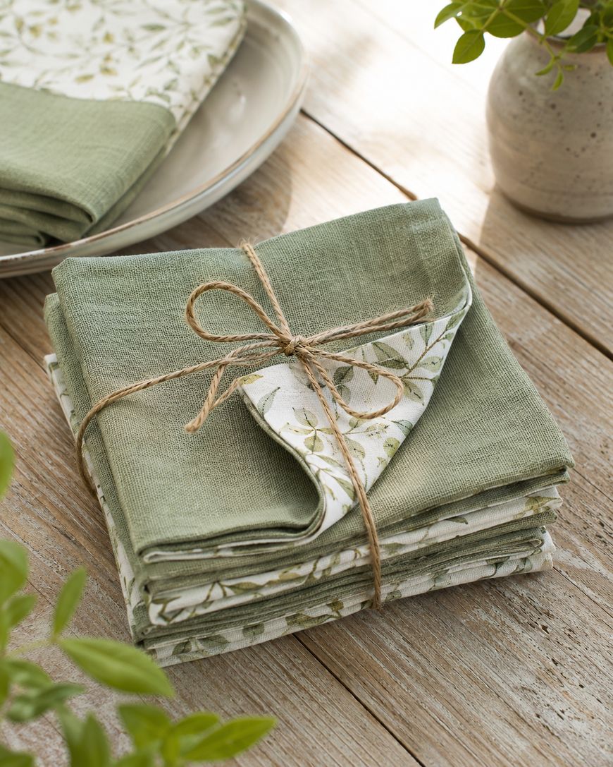 A stack of green and white patterned cloth napkins tied with twine sits on a rustic wooden table, with plates and potted plants nearby.