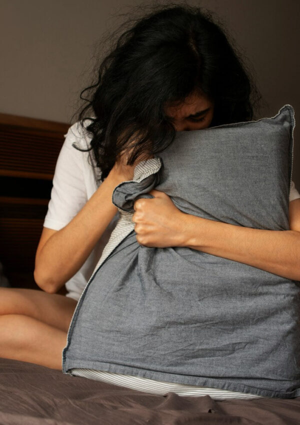 A person with dark, wavy hair sits on a bed, hugging a gray pillow tightly and pressing their face into it, suggesting feelings of sadness or distress.