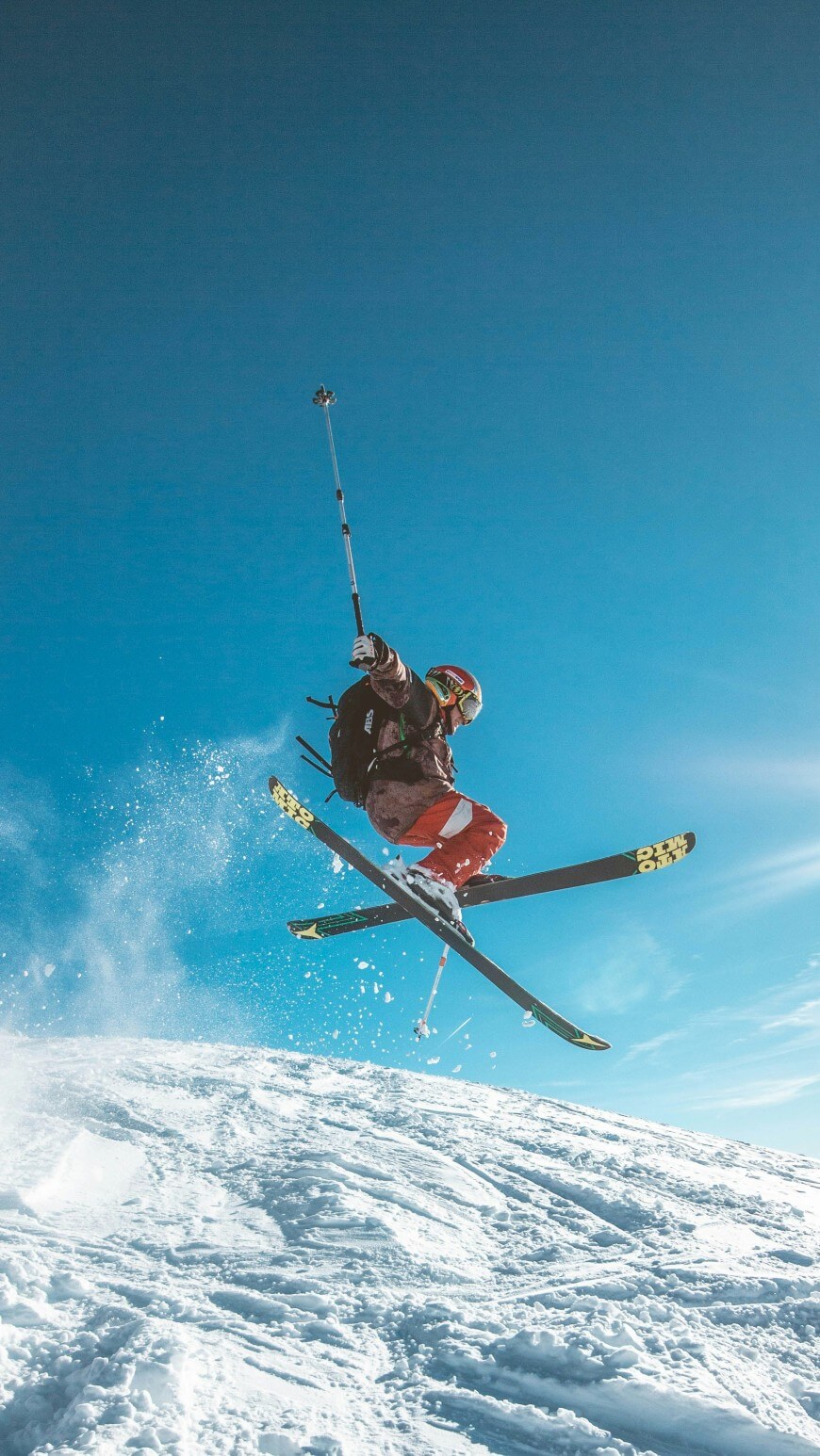 A skier in red pants and a helmet is mid-air performing a jump on a snowy slope under a clear blue sky, holding a ski pole out to the side.