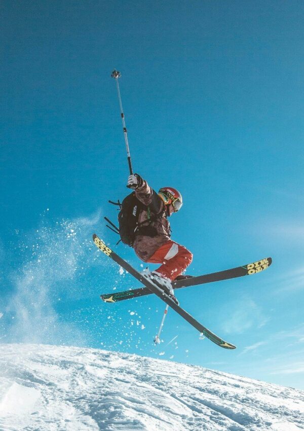 A skier in mid-air performs a jump on a snowy slope, holding ski poles and wearing a helmet, goggles, and winter gear against a clear blue sky.