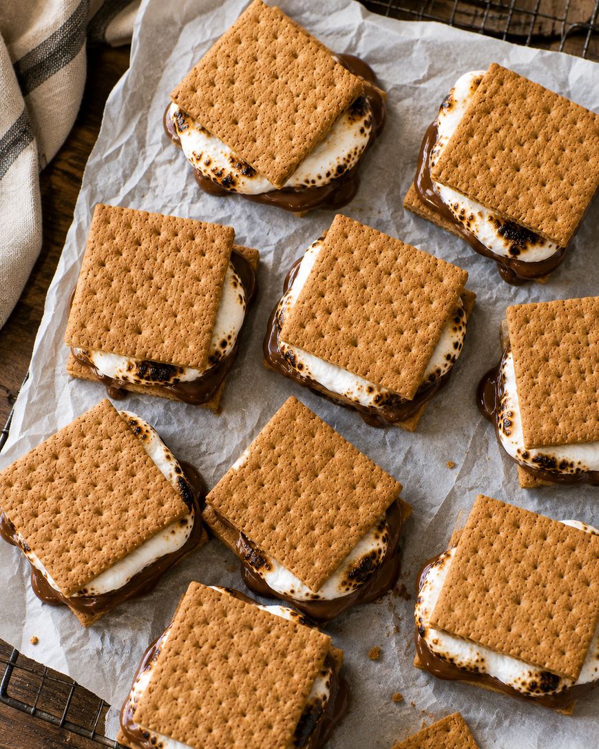 Nine s’mores made with toasted marshmallow and melted chocolate between square graham crackers are arranged on parchment paper on a cooling rack.