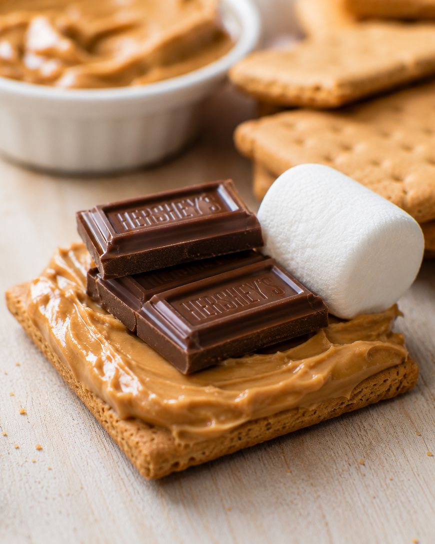 A graham cracker with peanut butter, topped with two pieces of chocolate and a large marshmallow, sits on a wooden surface with more crackers and a bowl of peanut butter in the background.