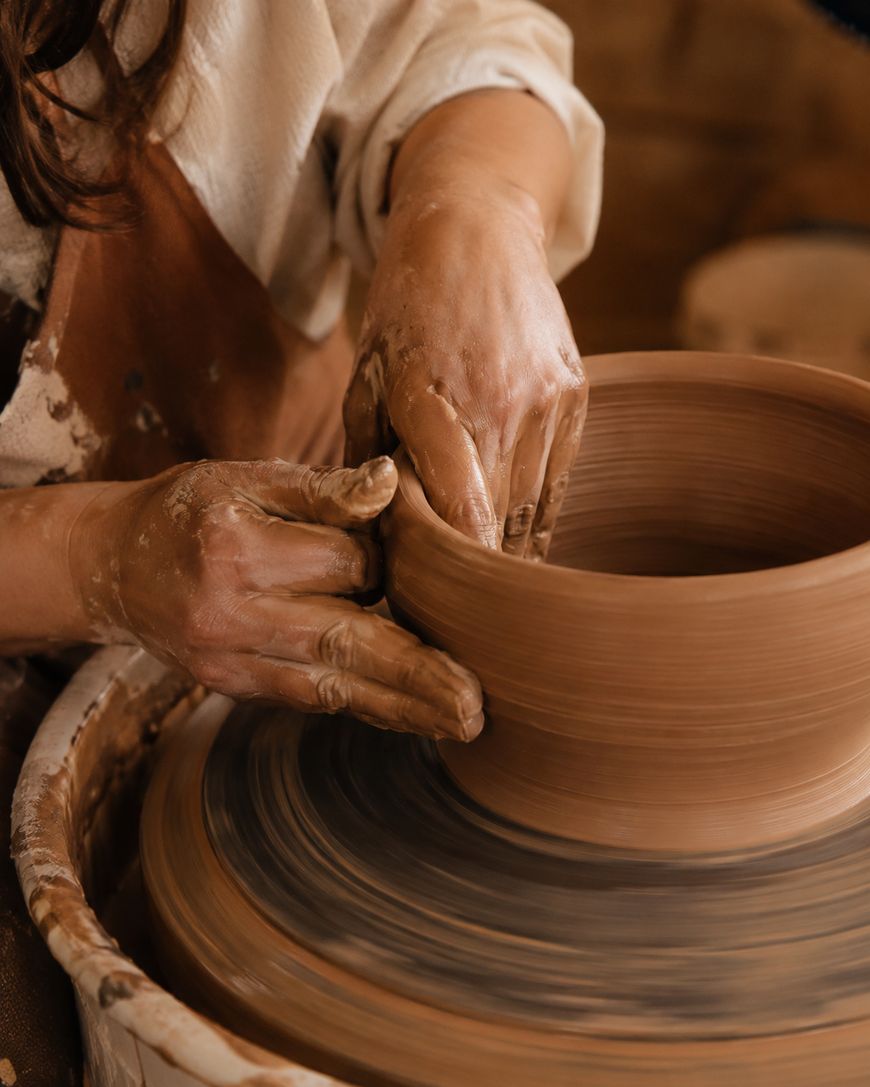Close-up of a person shaping a clay pot on a pottery wheel, with their hands covered in wet clay.