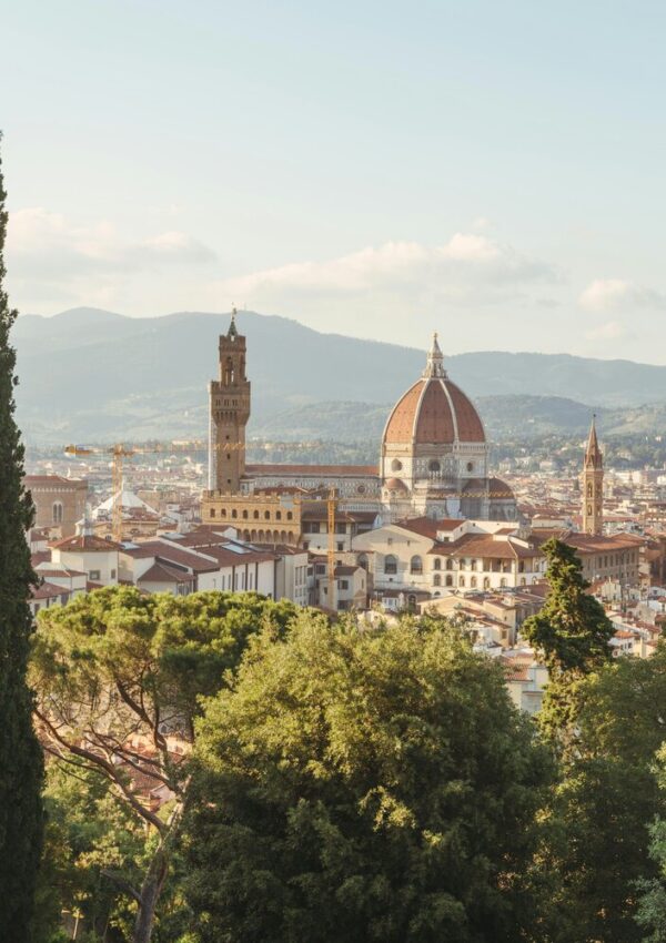 View of Florence, Italy with the Florence Cathedral’s dome, historic buildings, towers, and lush green trees in the foreground, set against distant hills under a partly cloudy sky—a perfect scene to inspire your stylish travel dreams as you explore Europe.
