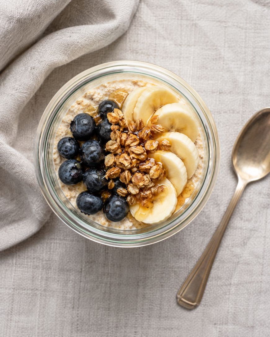 A glass bowl of oatmeal topped with sliced bananas, blueberries, granola, and honey sits on a textured cloth next to a spoon.