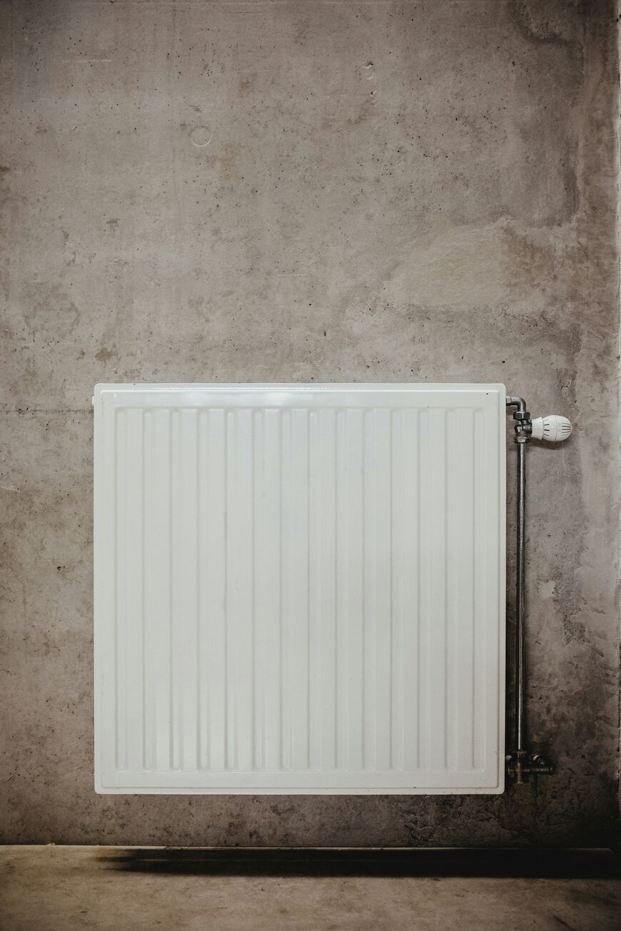 A white radiator attached to a gray concrete wall, with a temperature control valve on the right side and visible connecting pipes.