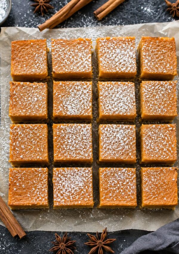 Sixteen pumpkin bars dusted with powdered sugar are arranged in a 4x4 grid on parchment paper. Cinnamon sticks and star anise are scattered around on a dark surface.