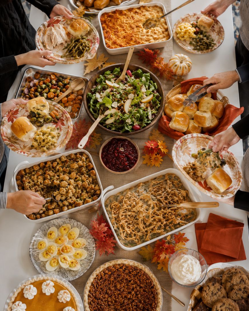 A Thanksgiving meal on a table with turkey, stuffing, green bean casserole, salad, mashed potatoes, deviled eggs, dinner rolls, cranberry sauce, sweet potato casserole, desserts, and hands serving food. Autumn leaves decorate the table.