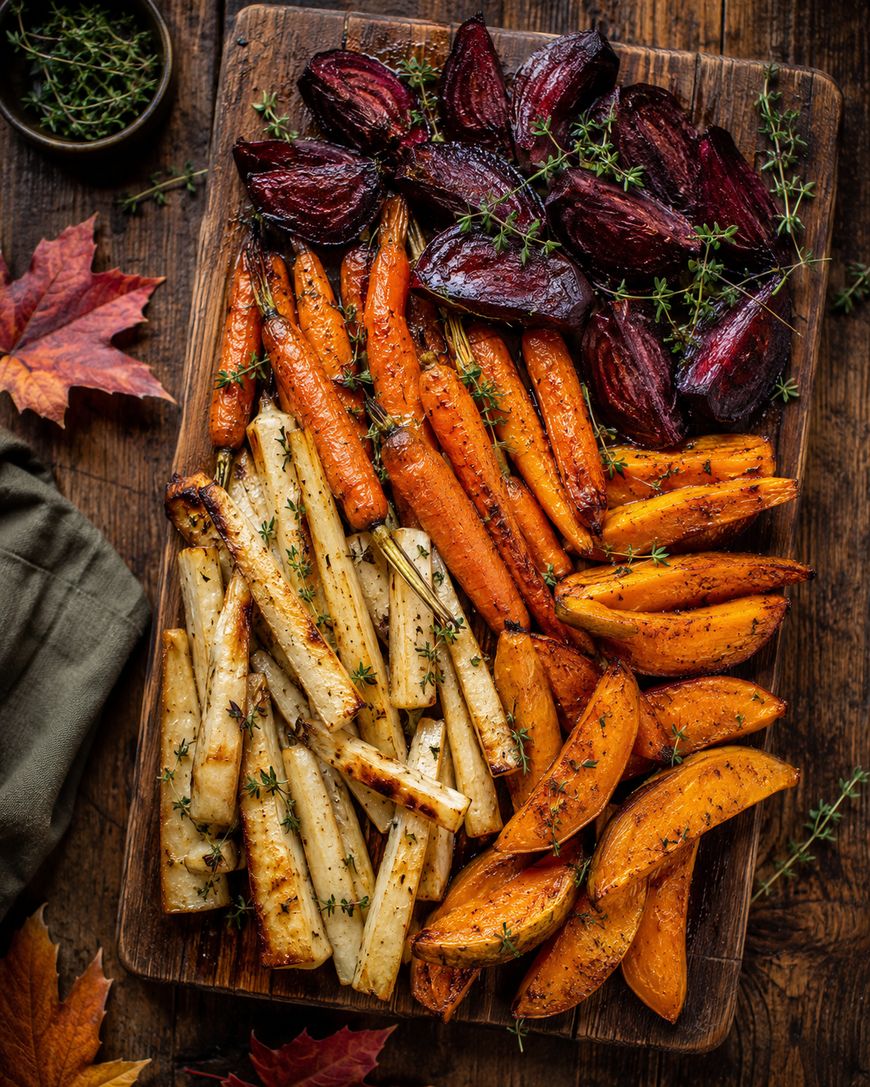 A wooden board with roasted beets, carrots, parsnips, and sweet potatoes, garnished with fresh herbs. Autumn leaves and a green napkin are nearby, creating a cozy, seasonal setting.
