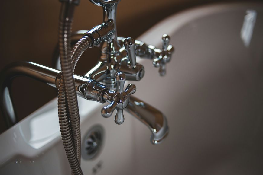 Close-up of a vintage-style chrome bathtub faucet with cross handles, attached to a white bathtub. A flexible shower hose is visible, along with the tub’s drain.