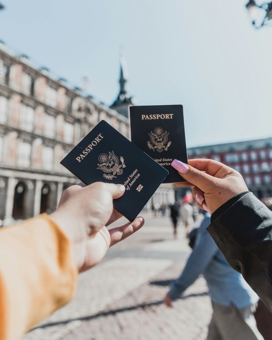 Two people holding U.S. passports up in a sunny, European-style plaza with blurred buildings and people in the background.