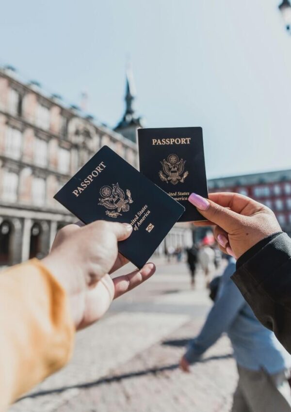 Two people holding up U.S. passports in front of a historic building in a sunlit plaza, with blurred pedestrians and architecture in the background.