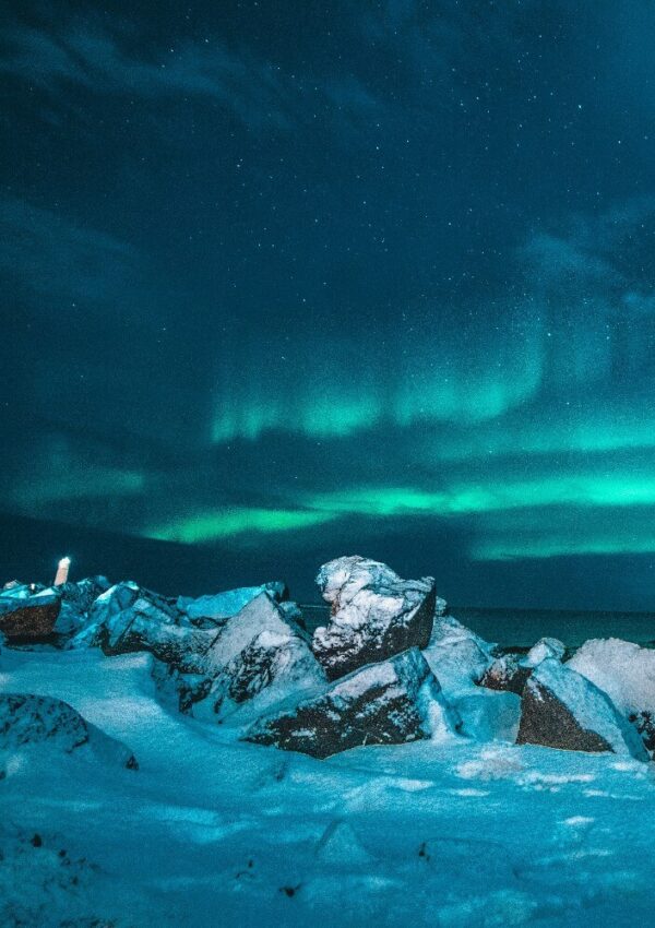 Snowy rocks line a cold shore under a starry night sky, illuminated by green northern lights. A small building glows faintly on the left, adding contrast to the icy, dramatic landscape.
