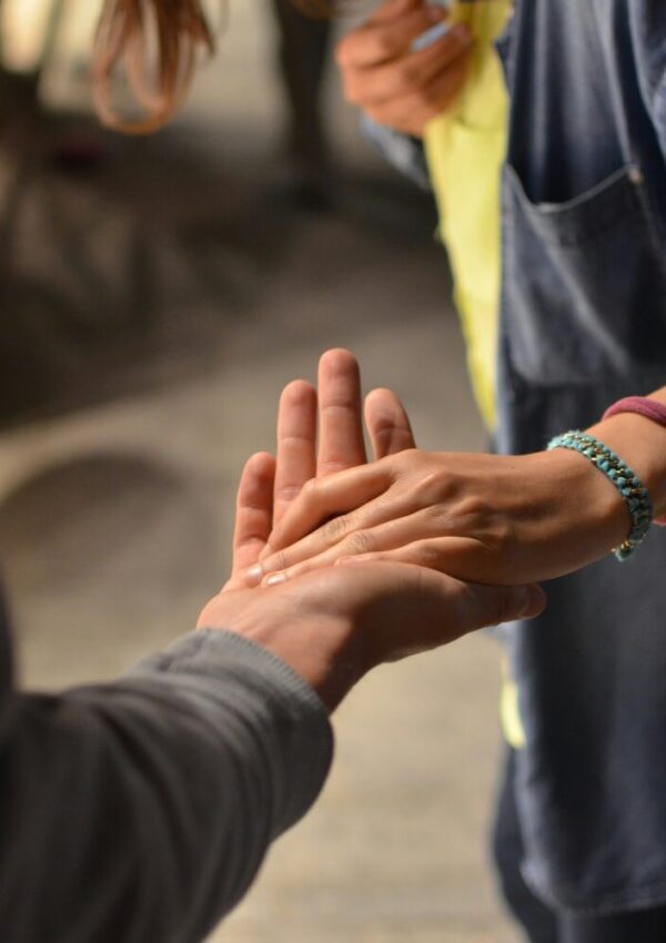 A close-up of two people gently touching hands, one person reaching out with their palm up while the other lightly places their hand on top. One person wears a blue shirt and a beaded bracelet.