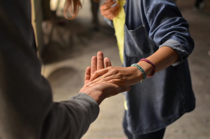 Two people holding hands; one person is gently touching the other’s open palm. Both are wearing casual clothing, and the background is softly blurred. The scene suggests comfort, support, or connection.