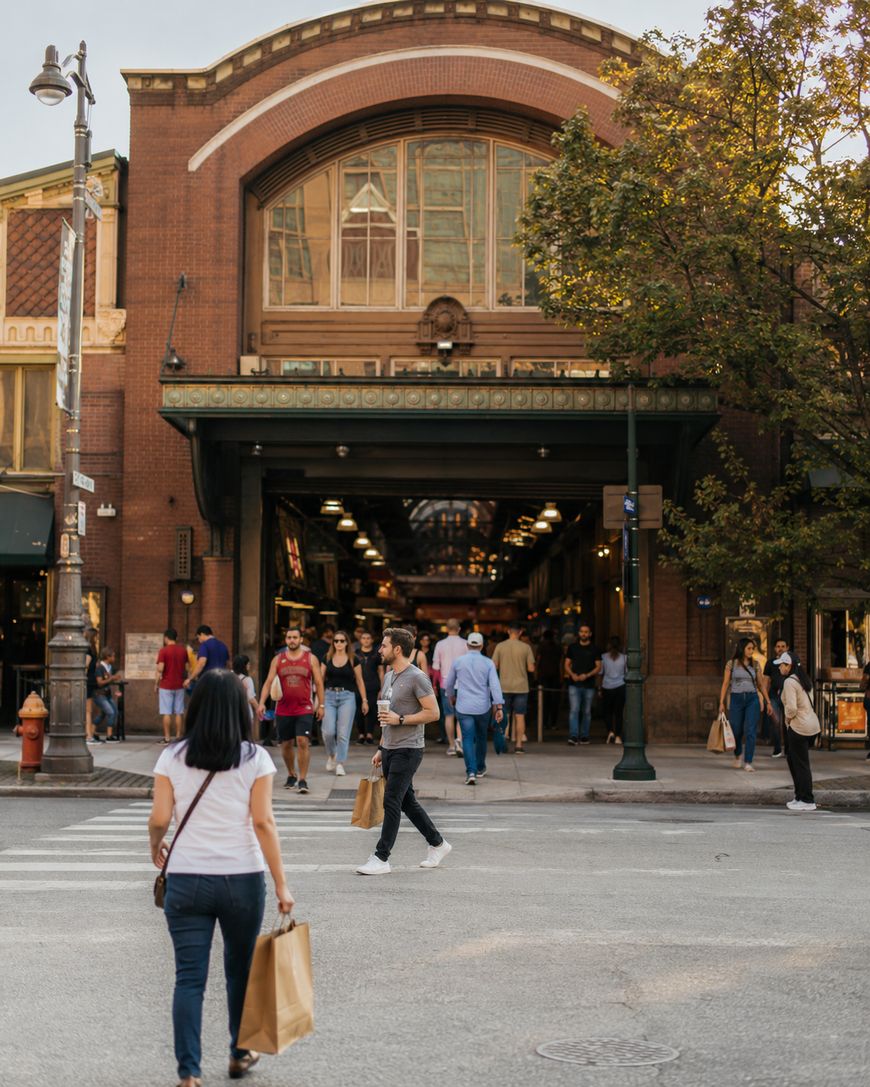 People with shopping bags cross the street toward a large, historic market building. Other pedestrians walk in and out of the entrance. Trees and street lamps line the road.