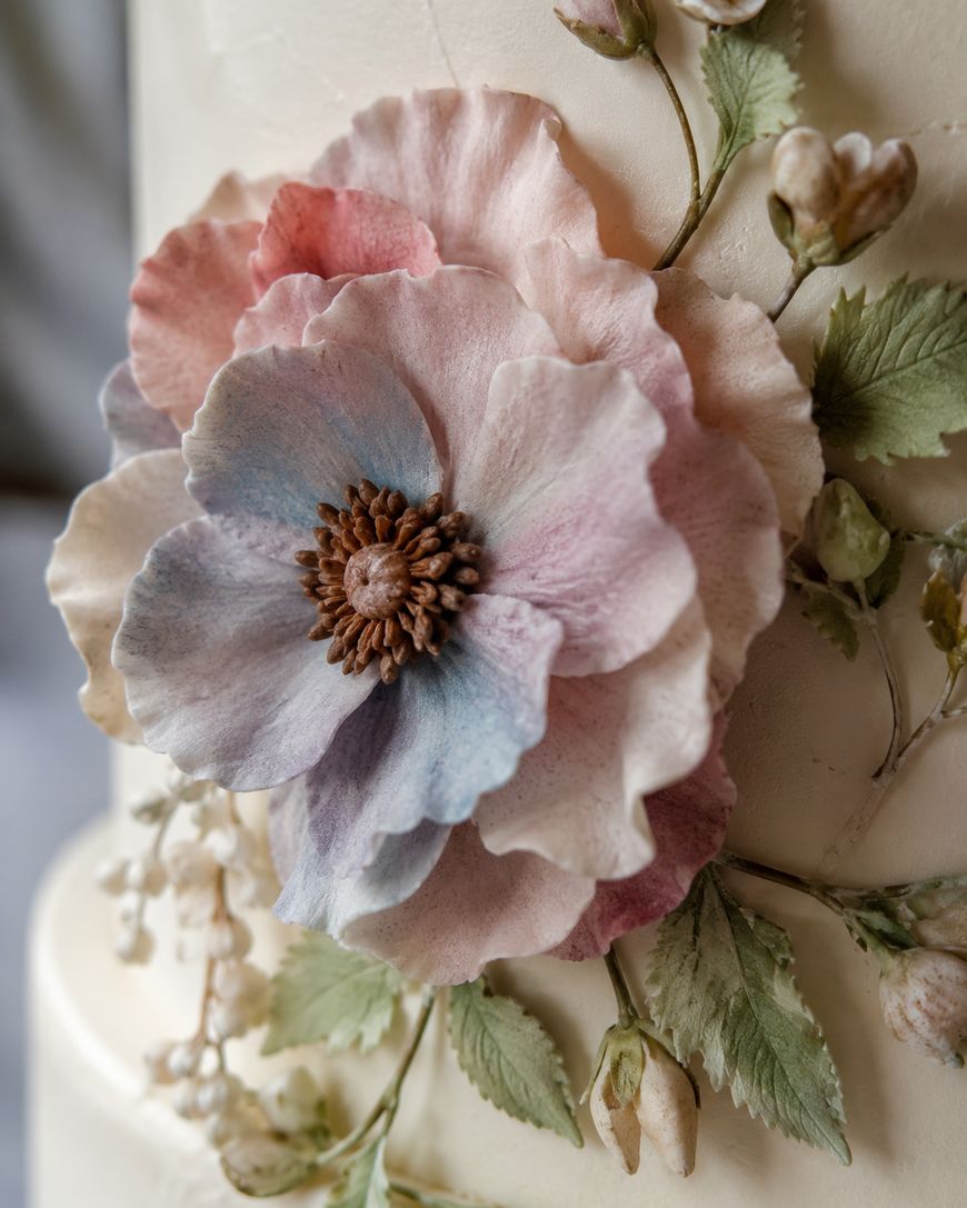 Close-up of a pastel-colored, realistic sugar flower decoration on a cake, with delicate petals in shades of pink, blue, and white surrounded by green leaves and small buds.