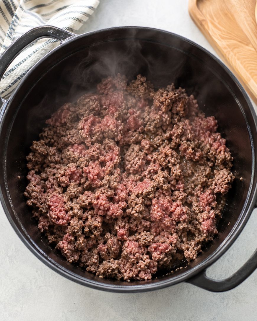 Ground beef is browning in a black pot, with some pink and some cooked brown pieces visible. Steam rises from the meat. A wooden board and striped towel are nearby on a light countertop.