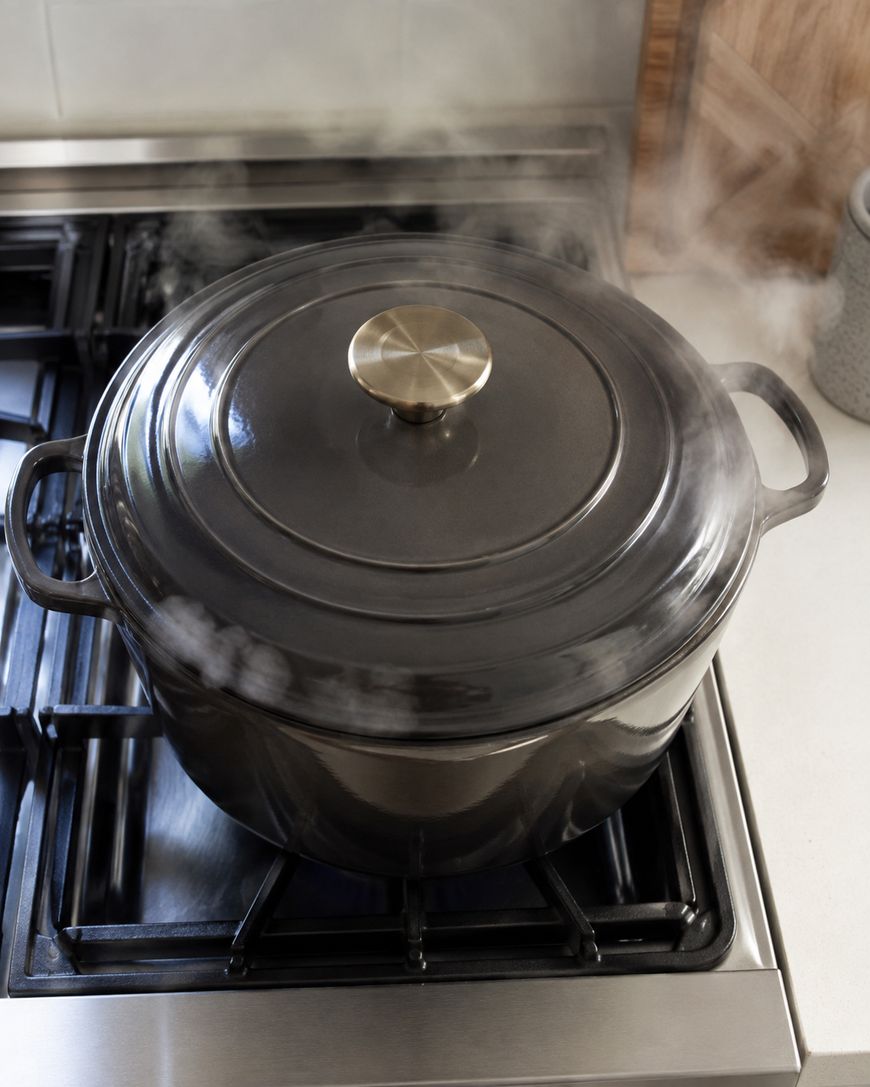 A black pot with a lid is steaming on a gas stove, with steam visible escaping around the edges of the lid. The stove is clean and the kitchen background is partially visible.