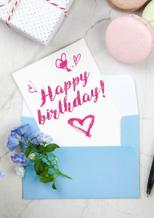 A Happy Birthday! card with pink lettering and hearts in a blue envelope, next to a pen, pastel macarons, blue flowers, and a wrapped gift on a white marble surface—perfect for long-distance celebrations that bring loved ones closer together.