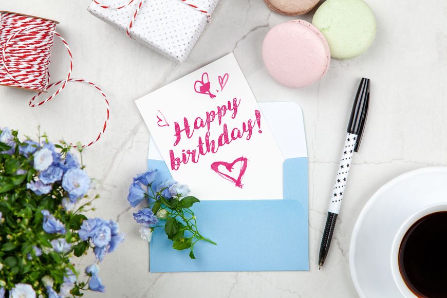 A birthday card with Happy birthday! in pink, flowers, a pen, macarons, twine, a wrapped gift, and a cup of coffee on a marble surface—perfect for long-distance celebrations or sending virtual birthday cards to loved ones far away.