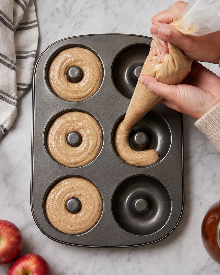 A person pipes batter into a six-cavity donut pan on a marble counter. A striped towel and apples are nearby.