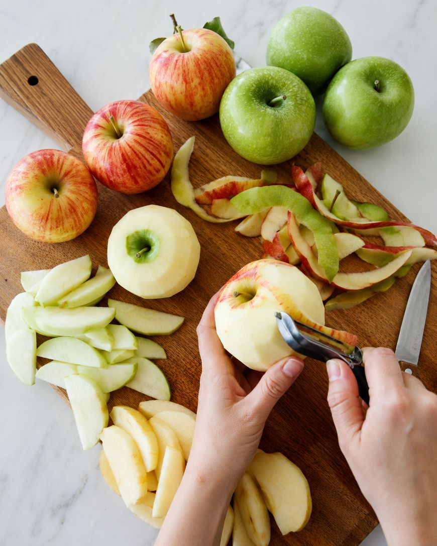 Hands peel a red apple on a wooden board with sliced apples, green and red whole apples, apple peels, and a knife on a white surface.