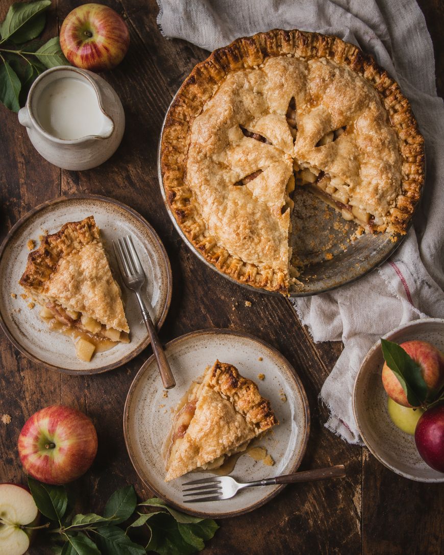 A golden apple pie with a slice missing sits on a wooden table. Two plates each hold a slice of pie, surrounded by apples, a pitcher of cream, and a linen napkin, creating a cozy, rustic scene.