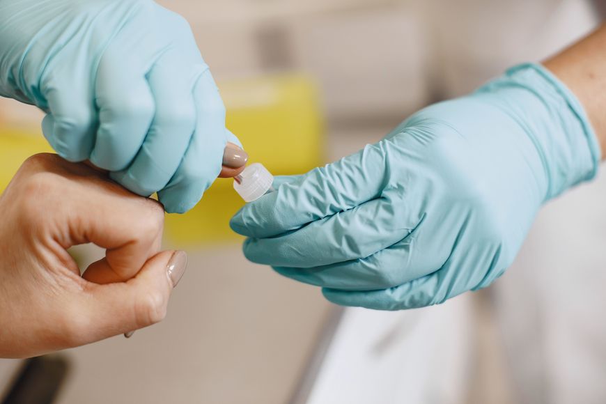 A person wearing blue gloves pricks another person’s fingertip with a small medical device, likely for a blood test or sample—demonstrating the accuracy often required in clinics and at home blood tests.