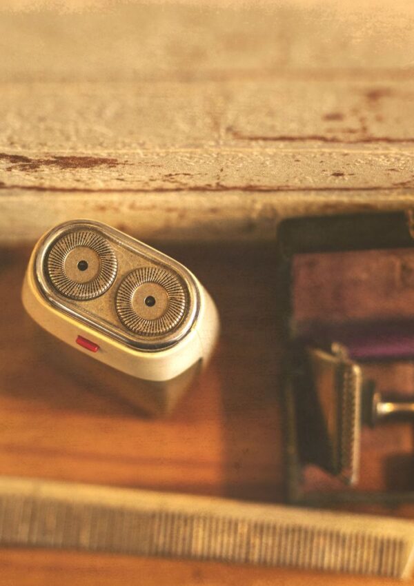 A close-up of an electric shaver with two circular blades, a comb, and part of a manual razor on a wooden surface near a worn white edge. Warm lighting gives a vintage feel.