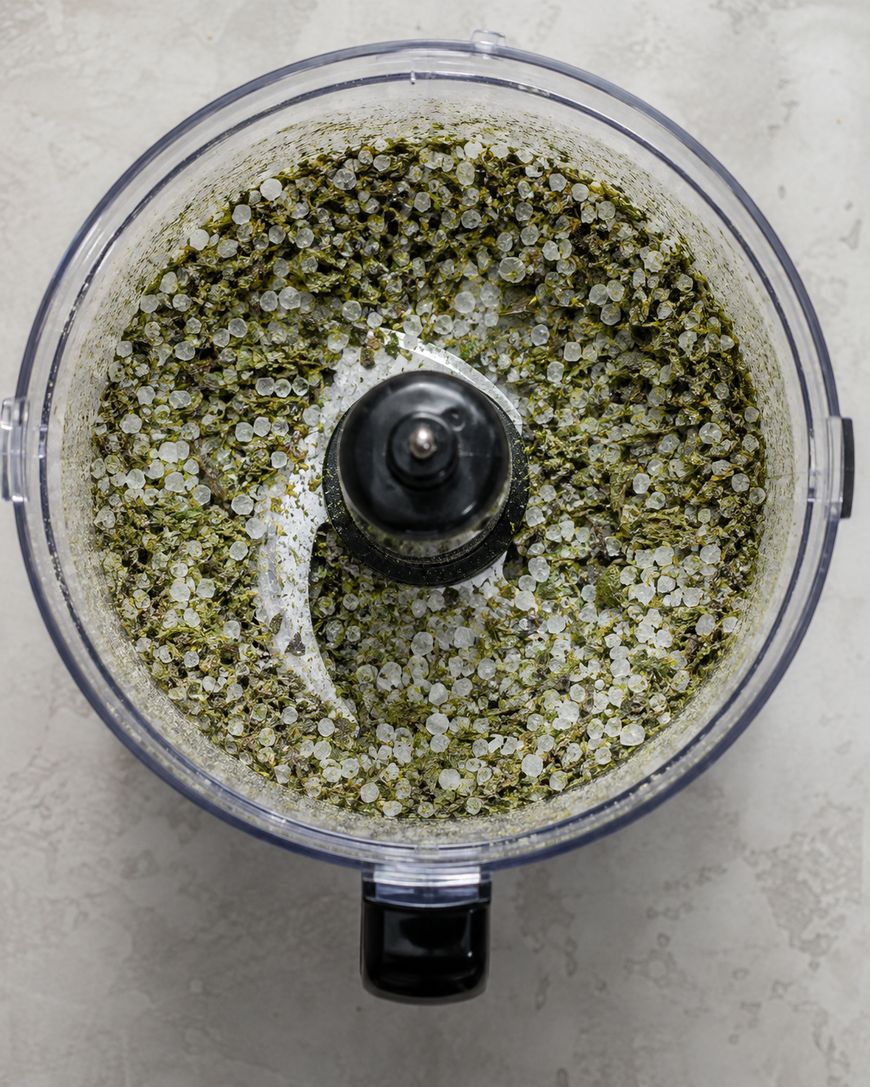 A top view of a food processor containing coarse sea salt mixed with green herbs, seen on a light textured surface.