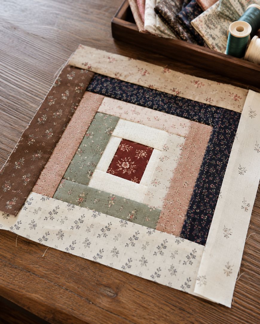 A quilt block made of patterned fabric strips in brown, cream, green, and navy is arranged in a square on a wooden table, with sewing supplies and stacked fabric nearby.