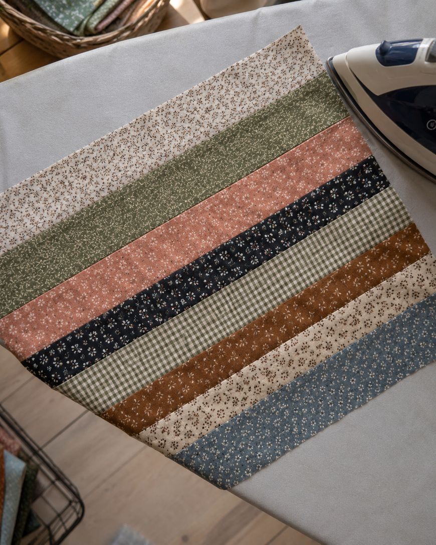 A close-up of a quilt block made from horizontal strips of patterned fabric on an ironing board, with an iron partially visible in the top right corner.