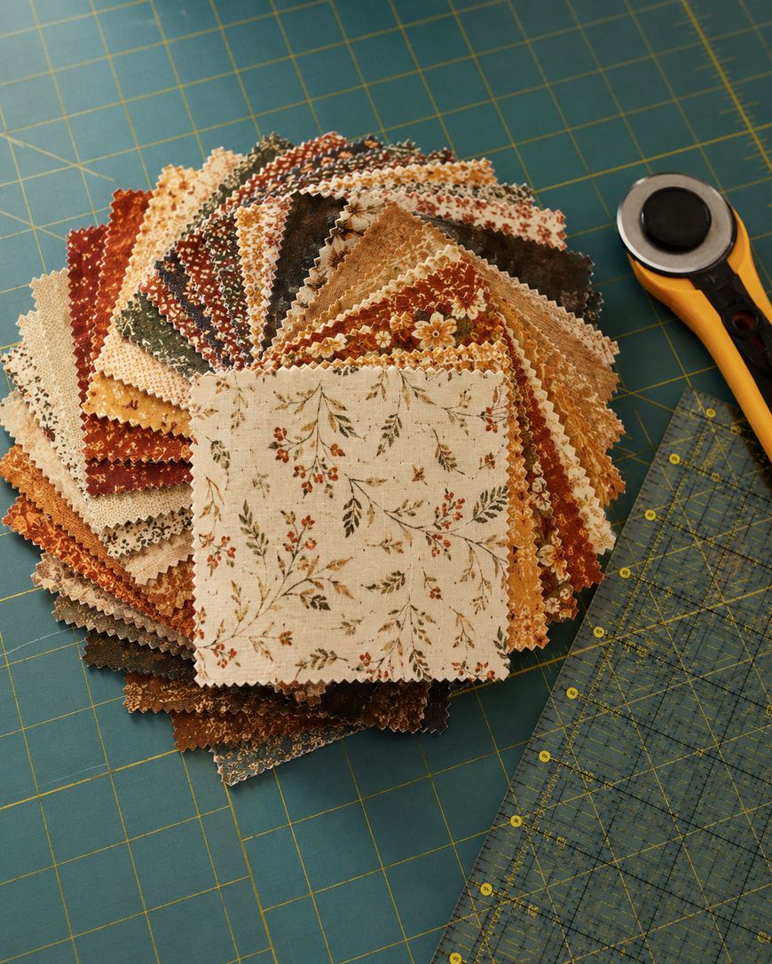 A stack of patterned fabric squares is fanned out on a green cutting mat beside a rotary cutter and a clear ruler, ready for quilting.