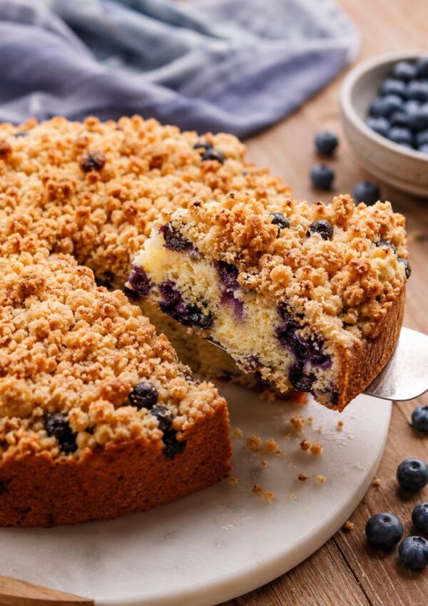 A slice of blueberry crumb cake is being lifted from the whole cake, which sits on a round cutting board. Fresh blueberries and a bowl of blueberries are nearby, and a blue cloth is in the background.