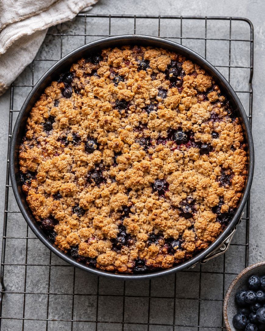 A round blueberry crumble cake in a baking pan sits on a cooling rack, with a bowl of fresh blueberries and a beige cloth nearby on a gray surface.
