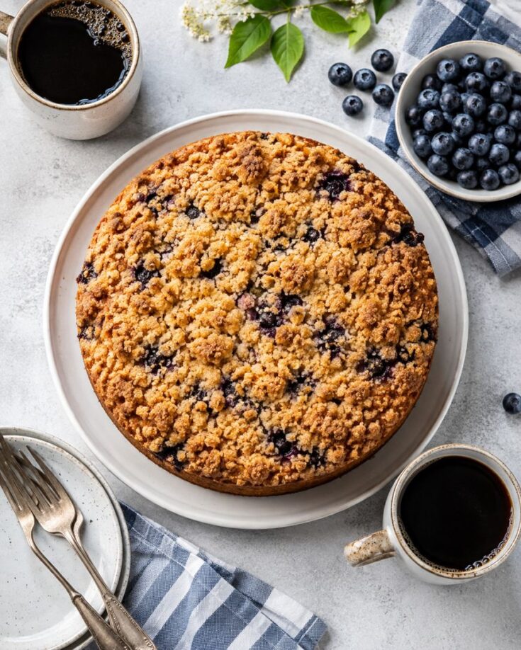 A round blueberry crumb cake on a white plate, surrounded by bowls of fresh blueberries, cups of black coffee, and plates with forks on a light table with a blue plaid napkin.
