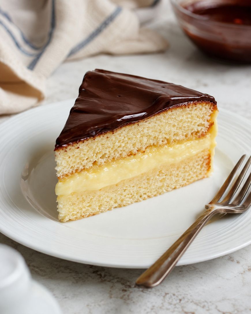 A slice of Boston cream pie with yellow custard filling and chocolate glaze sits on a white plate with a fork, with a napkin and bowl of chocolate in the background.