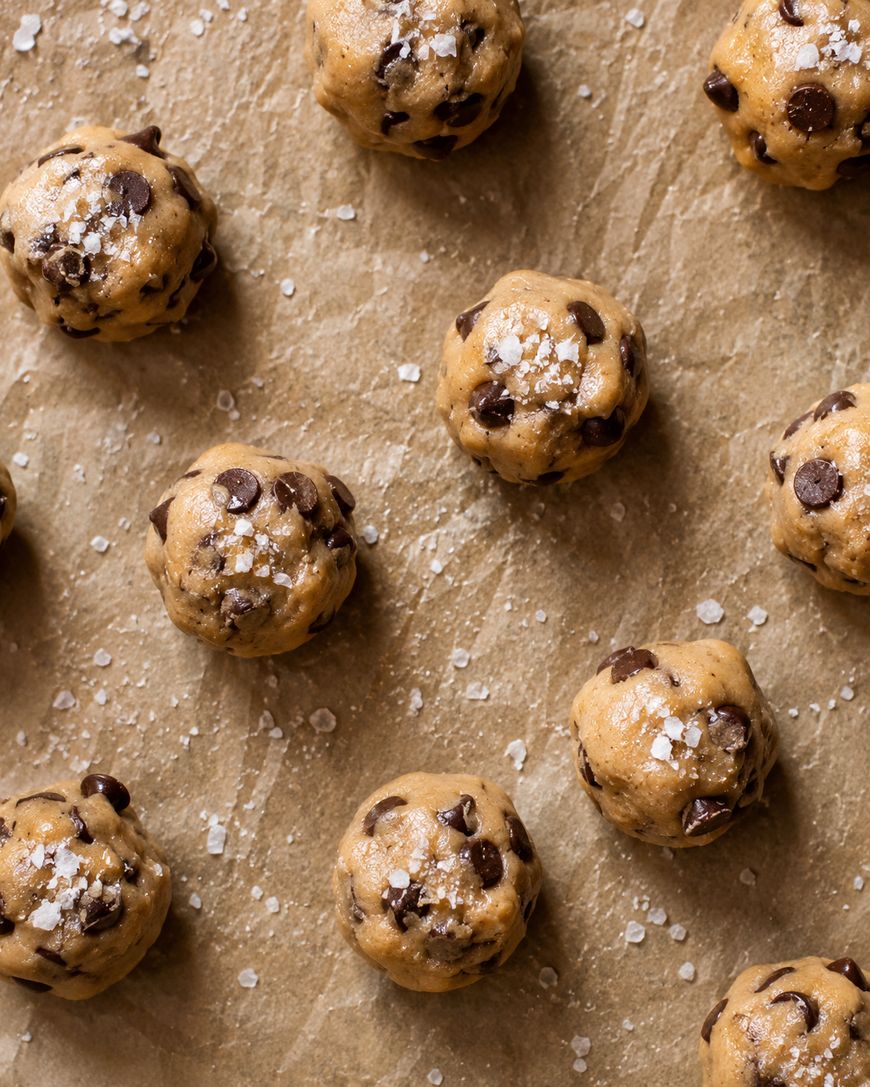 Balls of chocolate chip cookie dough sprinkled with coarse salt are arranged on a sheet of brown parchment paper, ready to be baked.