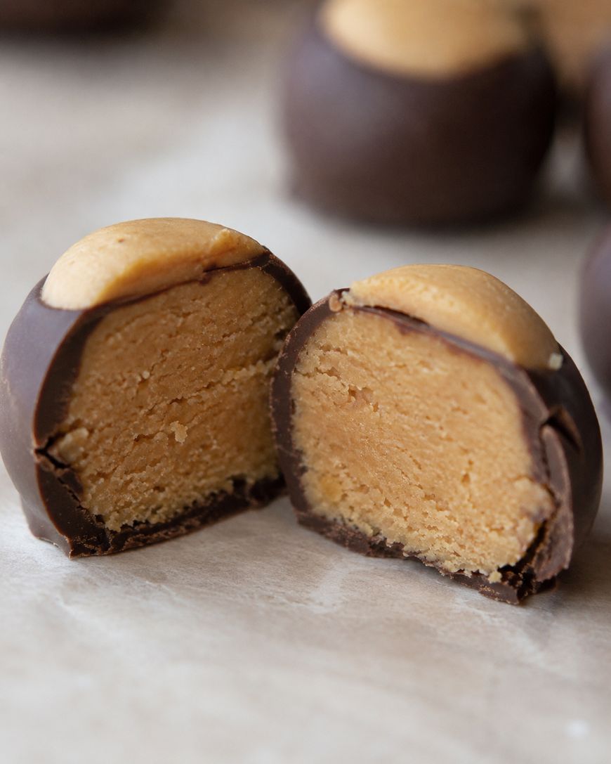 A close-up of a chocolate-coated peanut butter candy, with one piece cut in half to show the creamy peanut butter filling inside. Other candies are blurred in the background.