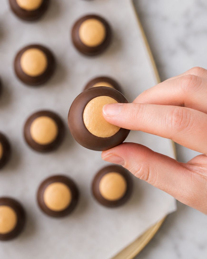 A hand holding a buckeye candy above a tray lined with parchment paper, with several more buckeye candies arranged in rows in the background.