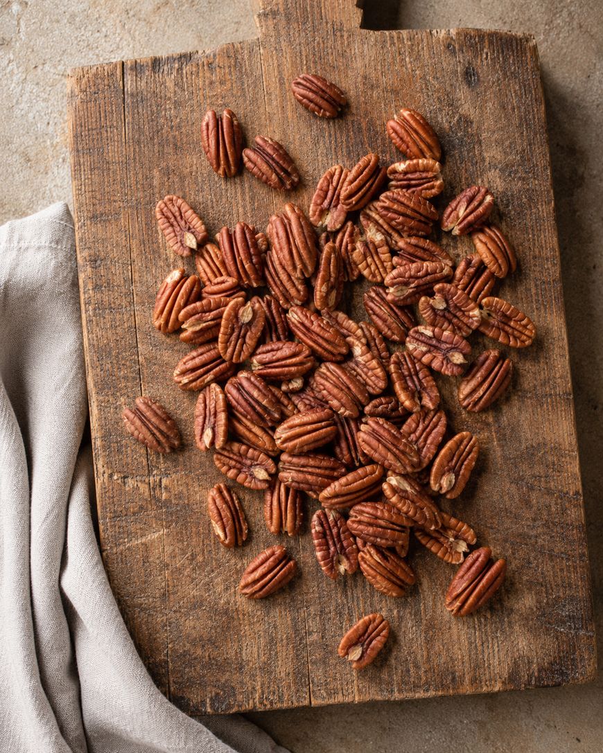 Pecan halves scattered on a rustic wooden cutting board, with a folded beige cloth next to the board on a stone surface.