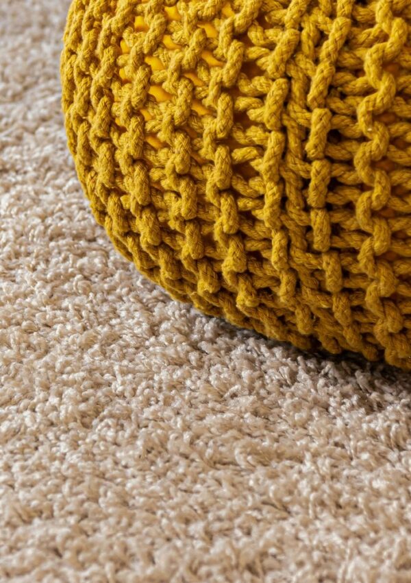 A close-up of a beige carpet with a yellow, chunky-knit pouf partially visible in the upper right corner.