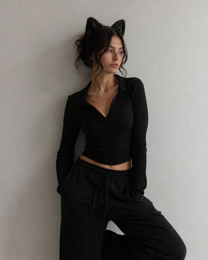 Woman with dark wavy hair wears black cat ear headband, a ribbed black top, and loose black pants, standing against a plain light-colored wall with hands in pockets, looking to the side.