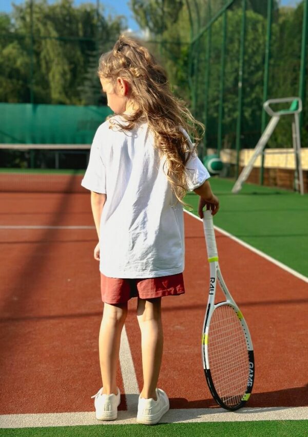 A young child with long hair stands on a tennis court, holding a tennis racket. The child faces away from the camera, wearing a white T-shirt, red shorts, and white sneakers.
