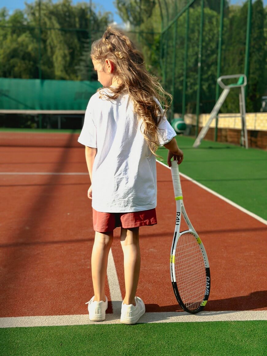 A young girl with long hair stands on a tennis court, holding a tennis racket by her side. She wears a white t-shirt, red shorts, and white sneakers, facing away from the camera.