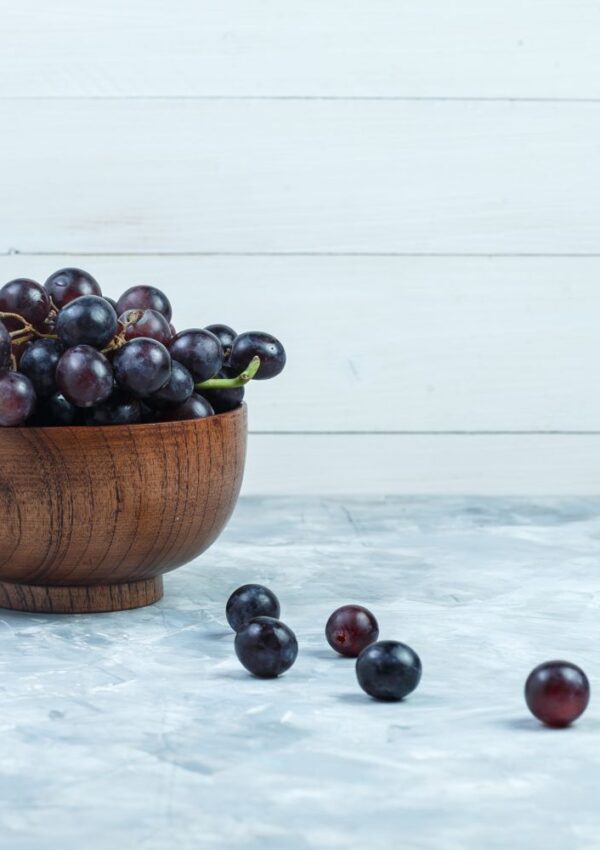 A wooden bowl filled with dark purple grapes sits on a gray textured surface, with several loose grapes scattered in front. A white wooden wall is in the background.